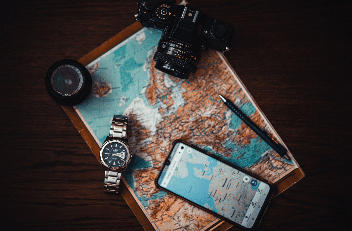 Vista cenital de una mesa. Sobre ella un mapa de Europa, una cámara de fotos con otro objetivo, un reloj de pulsera, un bolígrafo y un móvil con un mapa de Alemania y alrededores en la pantalla.