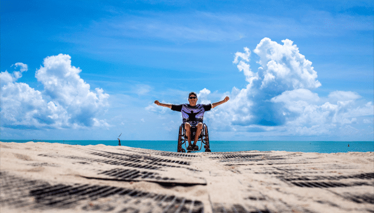En el centro de la imagen, un hombre en silla de ruedas y con gafas de sol, en una playa mirando a cámara, con el mar de fondo y un cielo soleado con alguna nube.