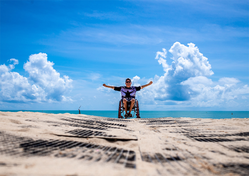En el centro de la imagen, un hombre en silla de ruedas y con gafas de sol, en una playa mirando a cámara, con el mar de fondo y un cielo soleado con alguna nube.