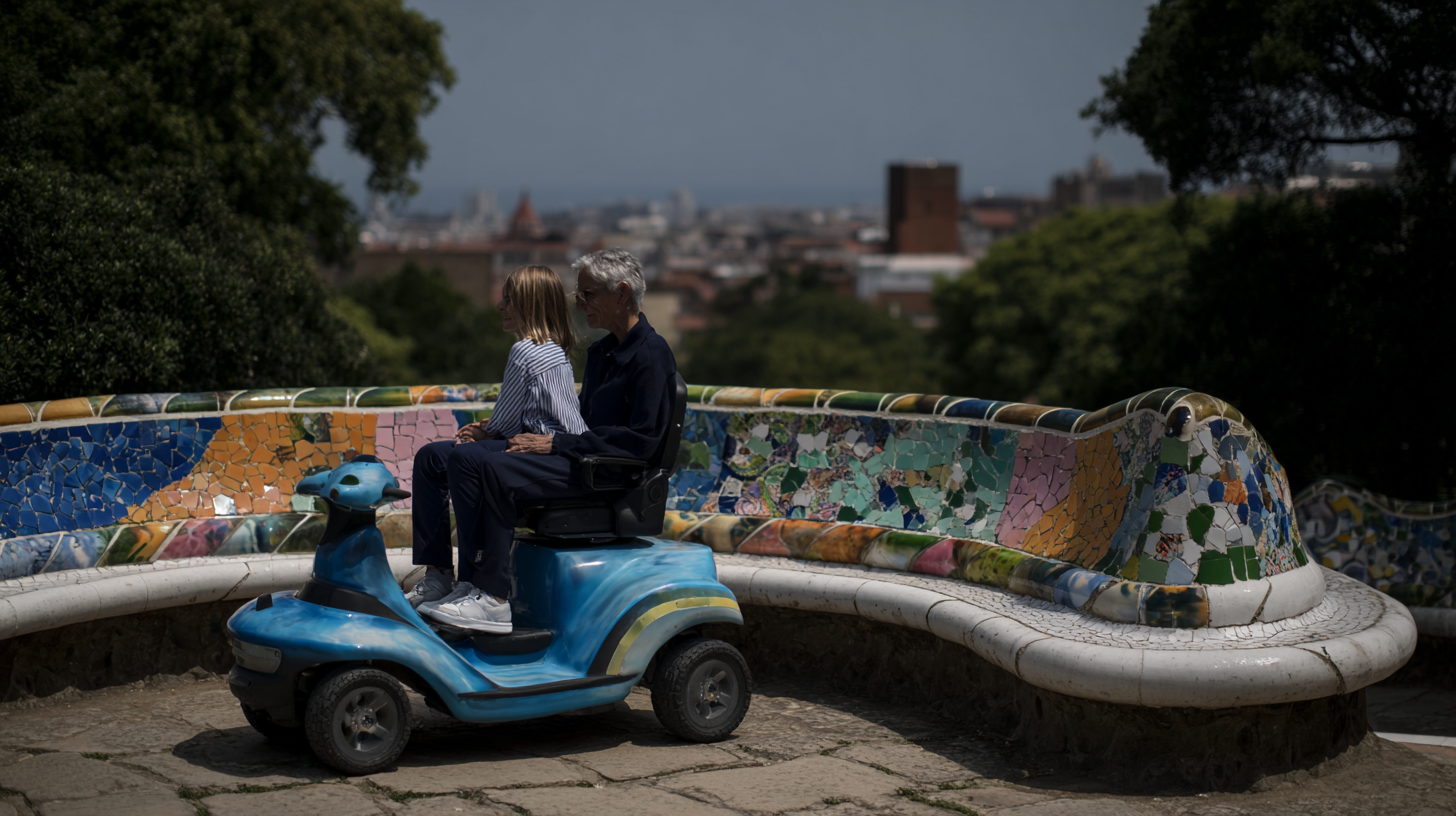 A father with a disability sitting on a mobility scooter for reduced mobility, holding his young daughter on his lap, accompanied by friends at Park Güell in Barcelona, enjoying an inclusive and wheelchair-accessible travel experience.